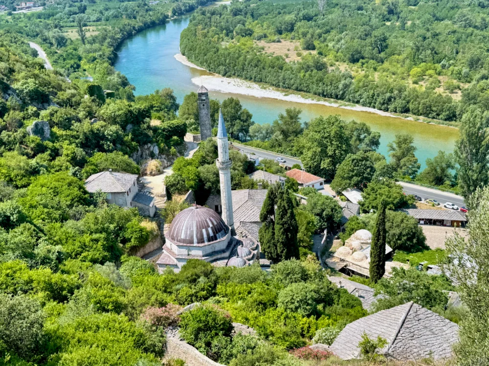 A historic hillside village in Bosnia and Herzegovina — a domed mosque and minaret among stone rooftops, above a bend in the turquoise river below.