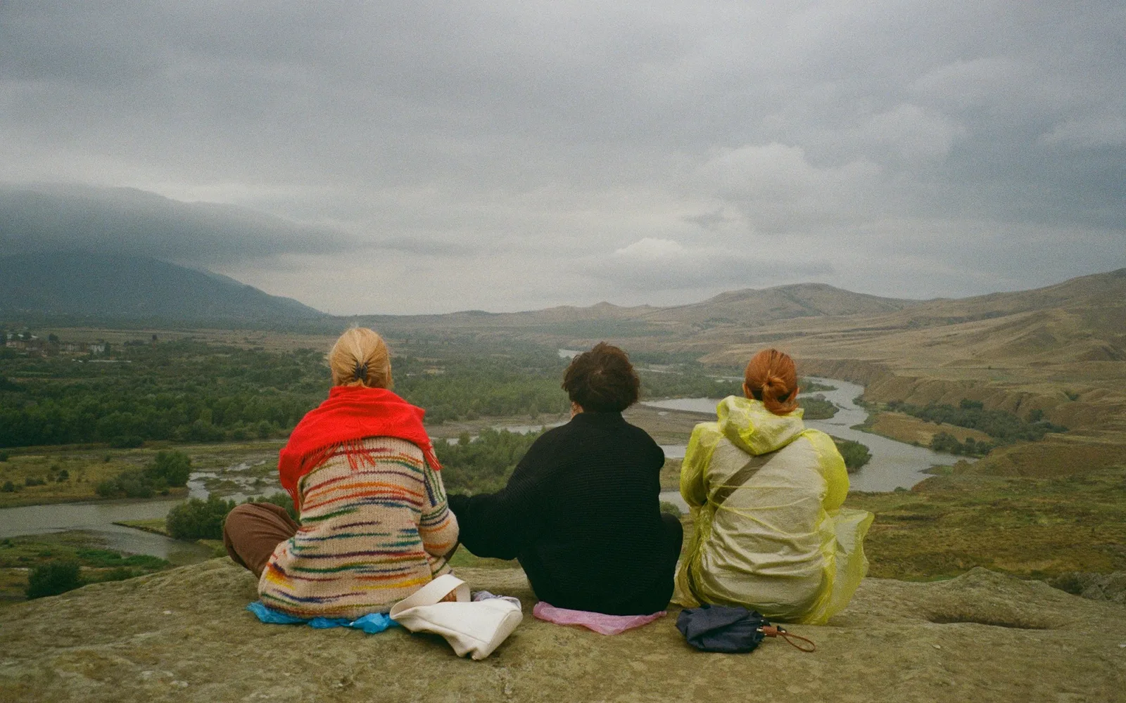 Three women sitting together on a rock, seen from behind, looking out over a river valley.